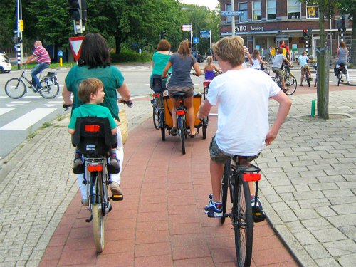 Dutch families on bikes on a cycle path at a junction. In the foreground we can see a teenage boy on a bike, next to him is his mother with the younger brother sat in a child seat on the back of her bike. Further away is another mother with her children in a box-bike.