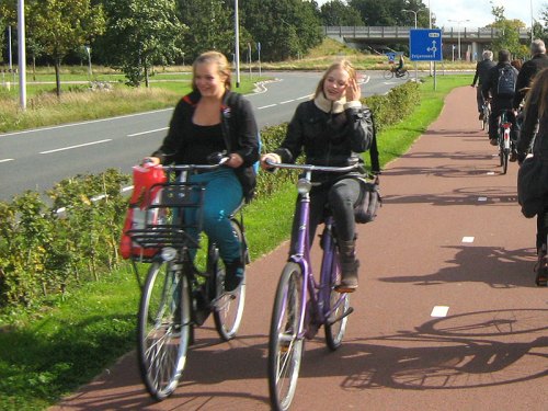Two teenagers ride their bikes on a cyclepath in the Netherlands, protected from the main road.