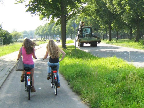 Two girls ride on a cycle path in Holland, beside a busy road with a tractor on it.