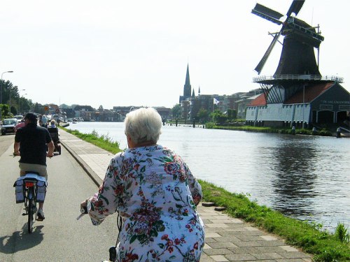 A grey-haired woman rides a bike alongside a canal and a windmill in the Netherlands