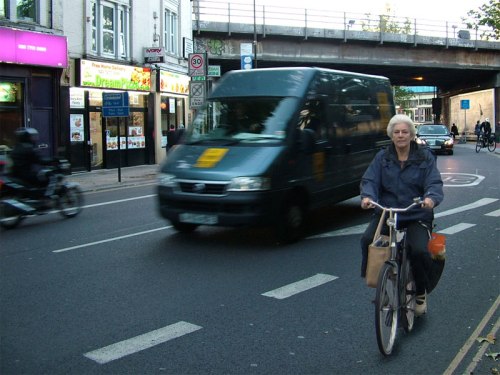 A photo of a Dutch woman riding a bike with shopping on it, juxtaposed with fast and dangerous on-road conditions in London.