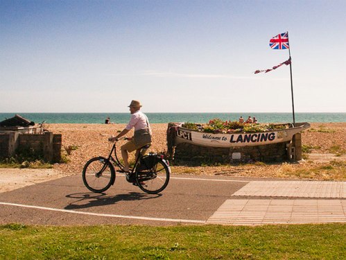 Mark Treasure riding on a motor-free cycle path alongside a beach, in bright clear weather. He is riding in casual clothes and wearing a trilby.