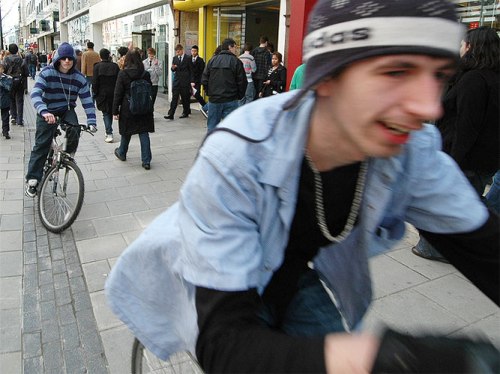 Two teenage boys ride aggressively along a busy footpath on a shopping street.