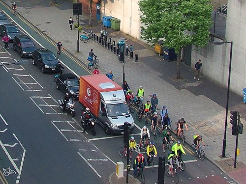 A birds' eye photo of a UK road junction with an ASL. All vehicles are positioned perfectly, no motor vehicles have entered the ASZ. But the ASZ can only hold around ten bikes, and there are twenty in shot, overflowing up the left-hand side of motor vehicles waiting.