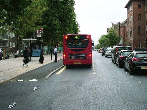 A photograph of a bus stopped on the new Royal College Street layout. There is room for cars to pass the stopped bus, but people boarding or alighting must stand in the cycle path while doing so. A bike user veers onto the footpath to avoid people getting on and off the bus.