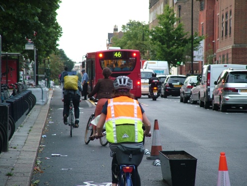 Another photograph of the same bus stop on Royal College Street. A taxi and a motorbike pass the stopped bus, while bike users and bus passengers are placed in conflict.