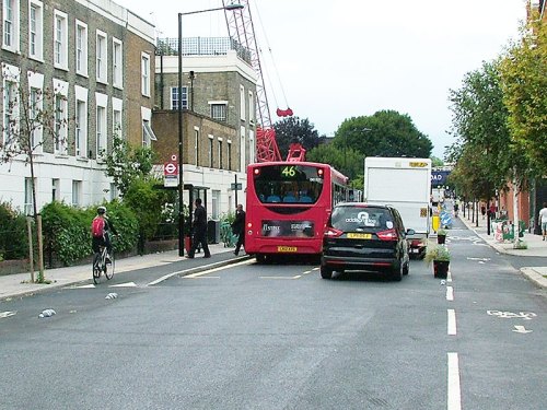 A photograph of the second bus stop on Royal College Street. There's enough space for a large van to pass the stopped bus, while a bike user has to stop while passengers board and alight the bus.