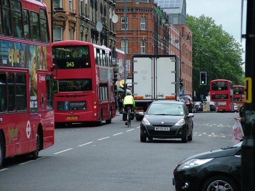 Kingsland High Street in Hackney, London. A bus is stopped, and a lorry is overtaking it. A cyclist dressed in high-visibility clothing follows the lorry, and a bus follows the cyclist.