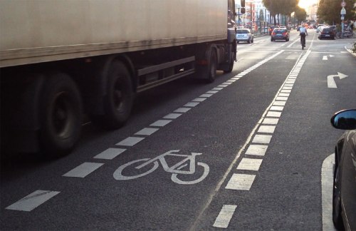 Photograph of Danziger Strasse in Berlin, showing a painted cycle lane on the carriage-way side of the parked cars, with a lorry thundering past.