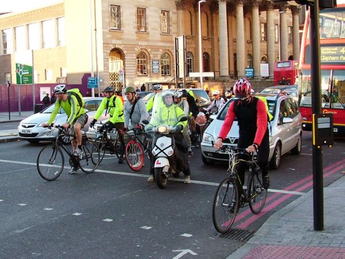 Cyclists in London waiting at traffic lights, surrounded by cars. All of the cyclists are wearing helmets, most are wearing hi-vis even though it's bright daylight.