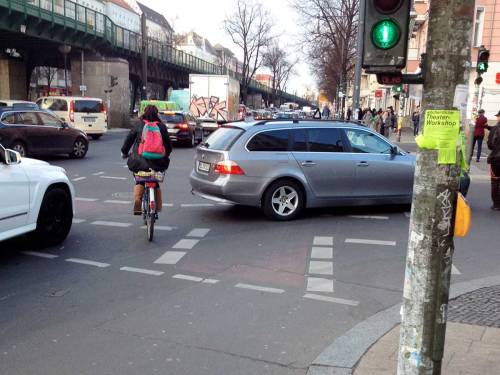 A traffic-signalled junction in Berlin, where turning cars must give way to pedestrians who have a green light, but the driver has blocked the cycle route due to poor design.