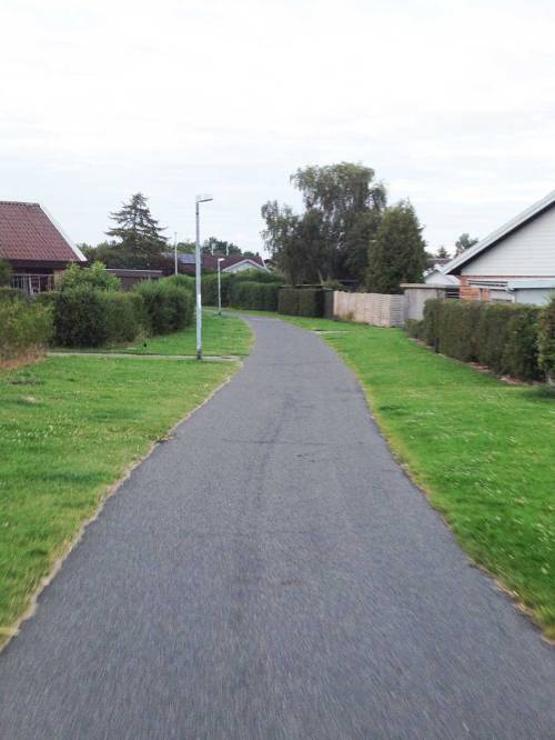A wide asphalt cycleway flows away the camera. Grass can be seen either side, with houses beyond it. There are street lights and side-paths connecting the houses.